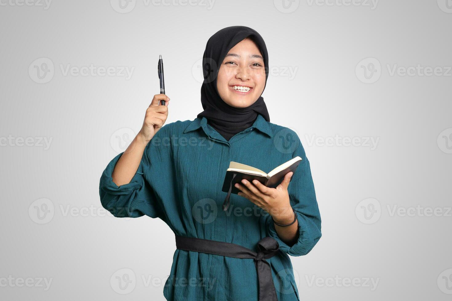 retrato de emocionado asiático musulmán mujer con hijab escritura en Nota libro y señalando arriba con bolígrafo. publicidad concepto. aislado imagen en blanco antecedentes foto