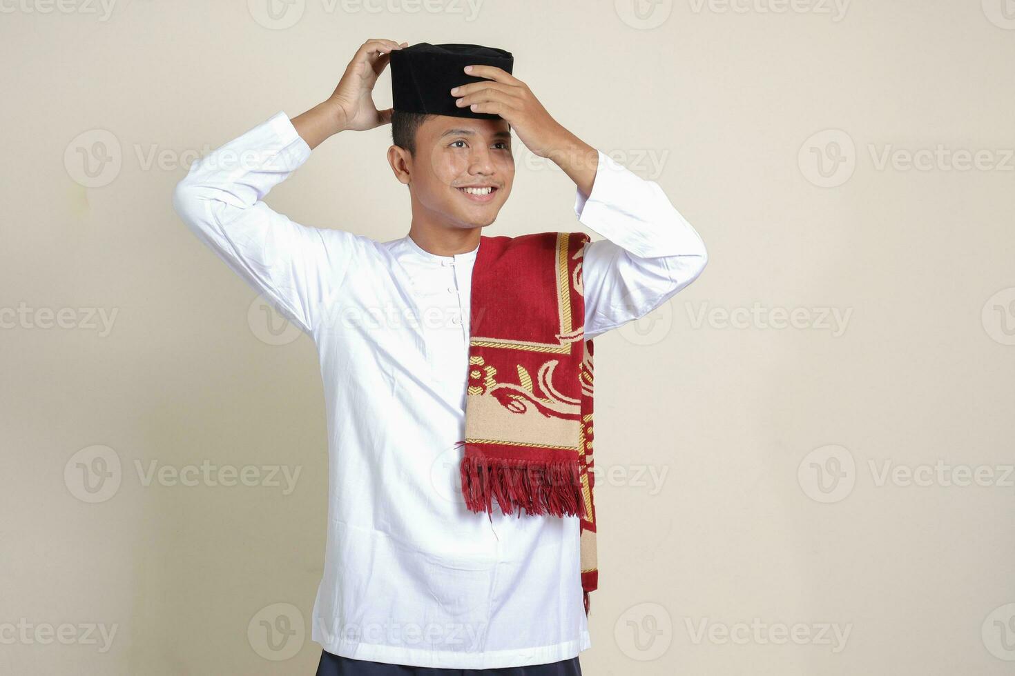 Portrait of attractive Asian muslim man in white shirt with skullcap trying to adjust his songkok or black skullcap. Isolated image on gray background photo