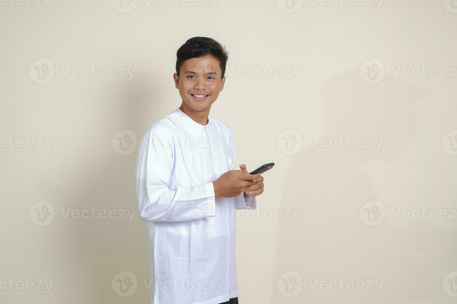 Portrait of attractive Asian muslim man in white shirt holding mobile phone with smiling expression on face. Advertising concept. Isolated image on gray photo