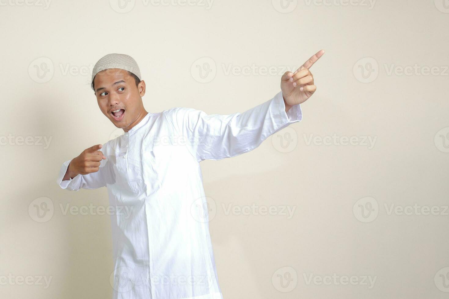 Portrait of attractive Asian muslim man in white shirt with skullcap showing product and pointing with his hand and finger to the side. Advertising concept. Isolated image on gray background photo