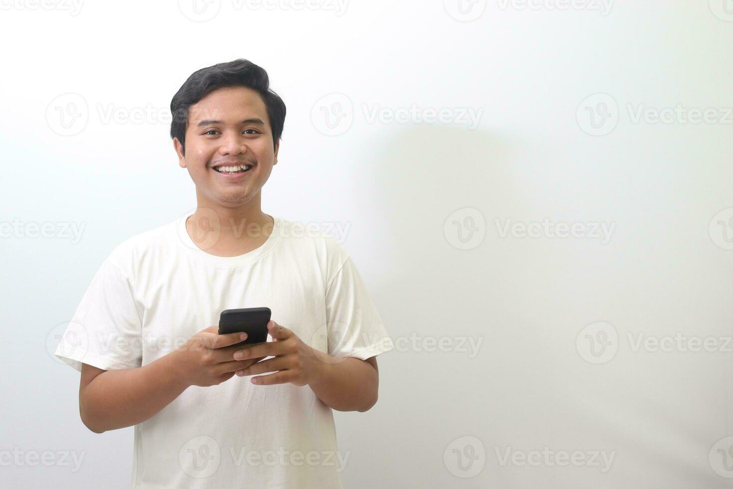 Portrait of shocked Asian man in white t-shirt standing against white background, holding a mobile phone while making surprise hand gesture. Wow face expression. Advertising concept. photo