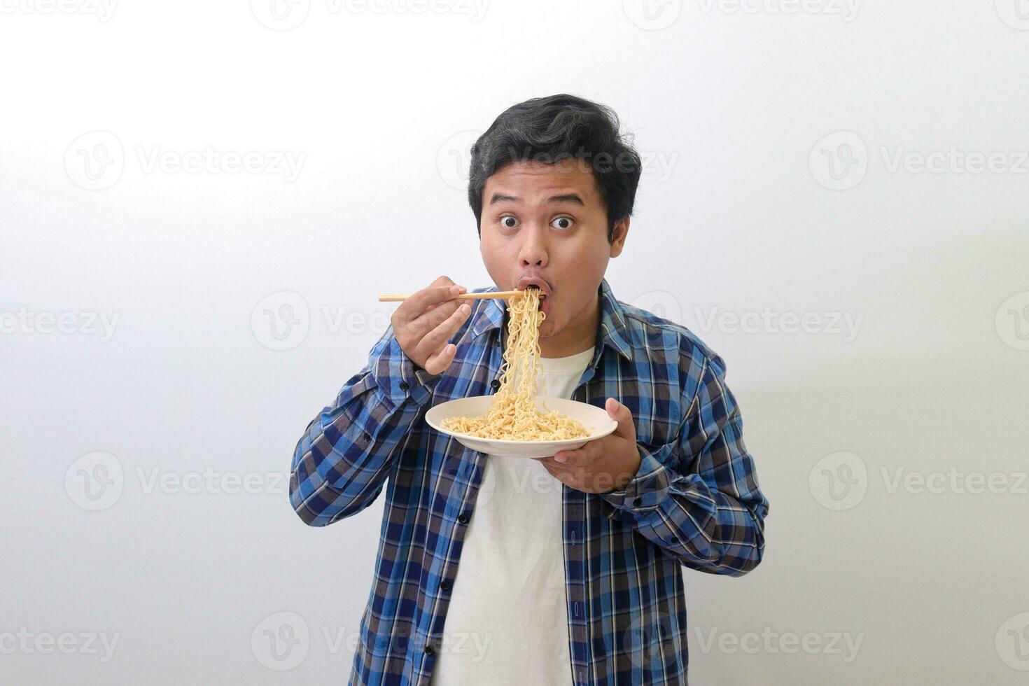 retrato de contento asiático hombre en azul tartán camisa comiendo delicioso instante tallarines con palillos servido en lámina. aislado imagen en blanco antecedentes foto