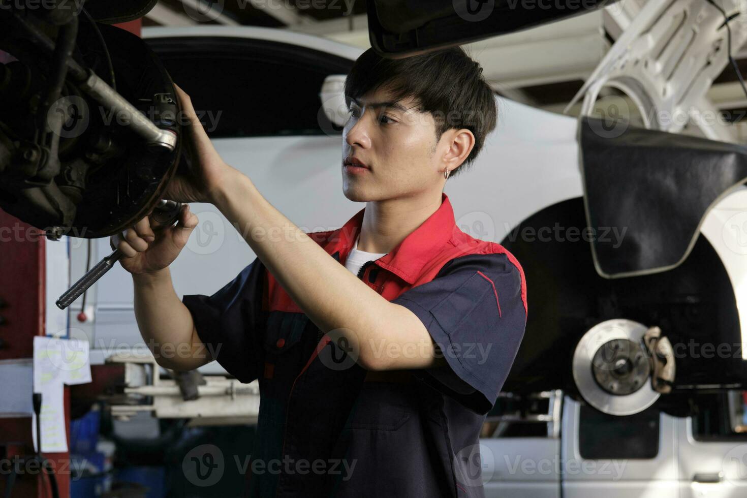 One young expert Asian male automotive mechanic technician is screwing car wheel nuts on lifting with wrench for repair at garage. Vehicle maintenance service works, industry occupation business jobs. photo