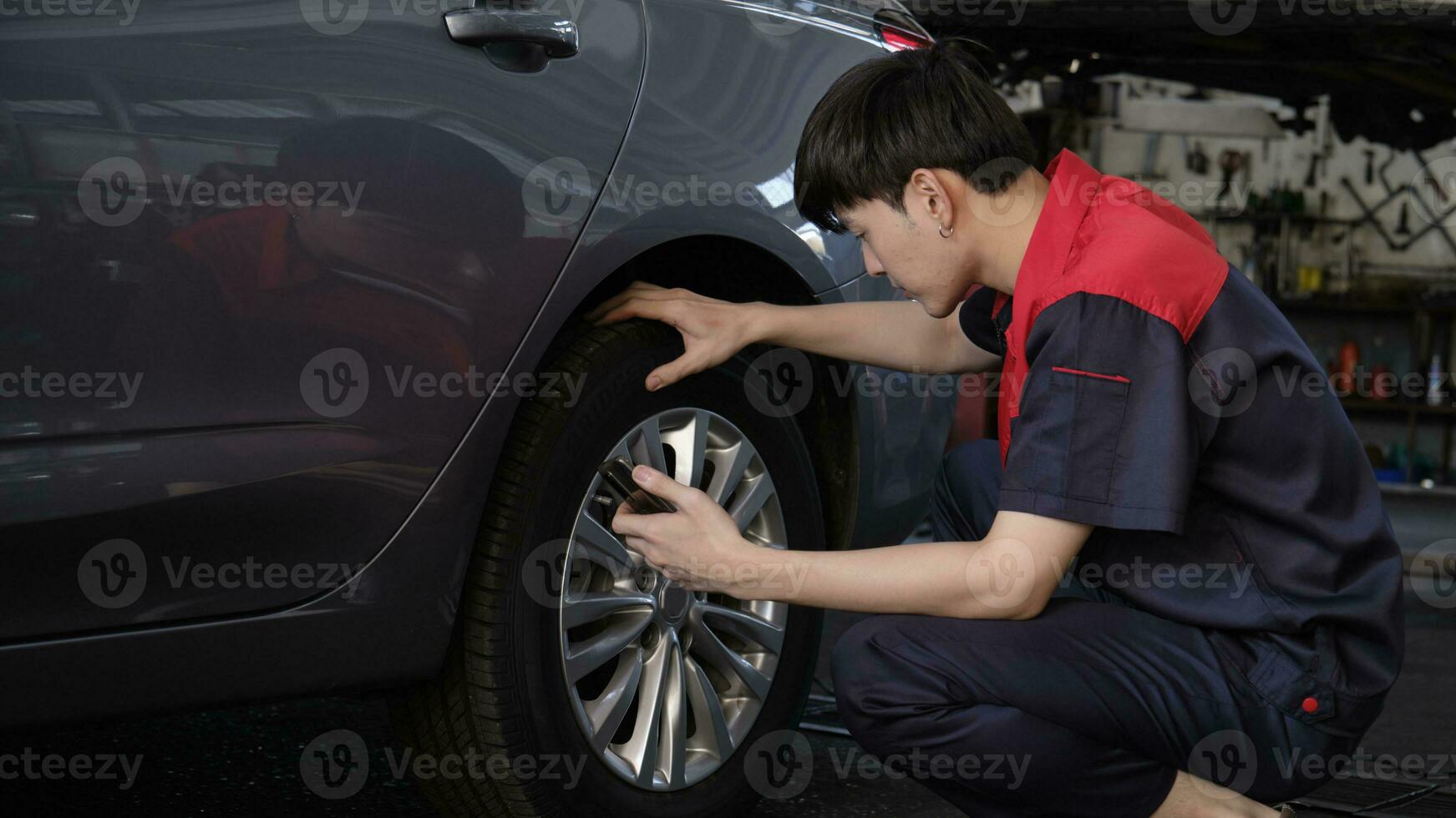 joven masculino asiático profesional automotor mecánico trabajador cheques neumático presión por teléfono inteligente solicitud a un coche cochera, experto en mantenimiento vehículo servicio, y fijación ocupaciones auto industria. foto