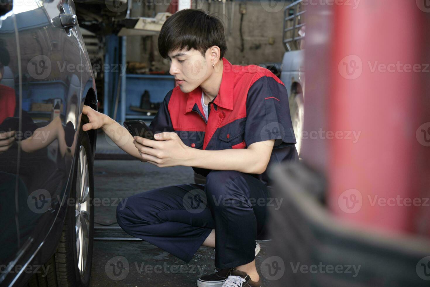 joven masculino asiático profesional automotor mecánico trabajador cheques neumático presión por teléfono inteligente solicitud a un coche cochera, experto en mantenimiento vehículo servicio, y fijación ocupaciones auto industria. foto