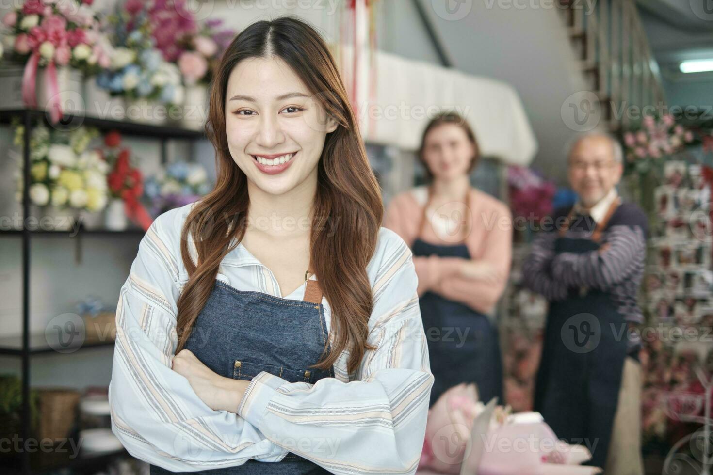 Beautiful young Asian female florist worker in apron, arms crossed, smiles and looks at camera in front of colleagues team in colorful flower shop, small business occupation, happy SME entrepreneur. photo