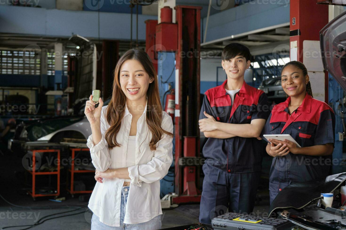 Young Asian female customer shows car key with automotive mechanic workers, happy smile in quality trust at maintenance garage, professional vehicle service center, fix check and repair auto industry. photo