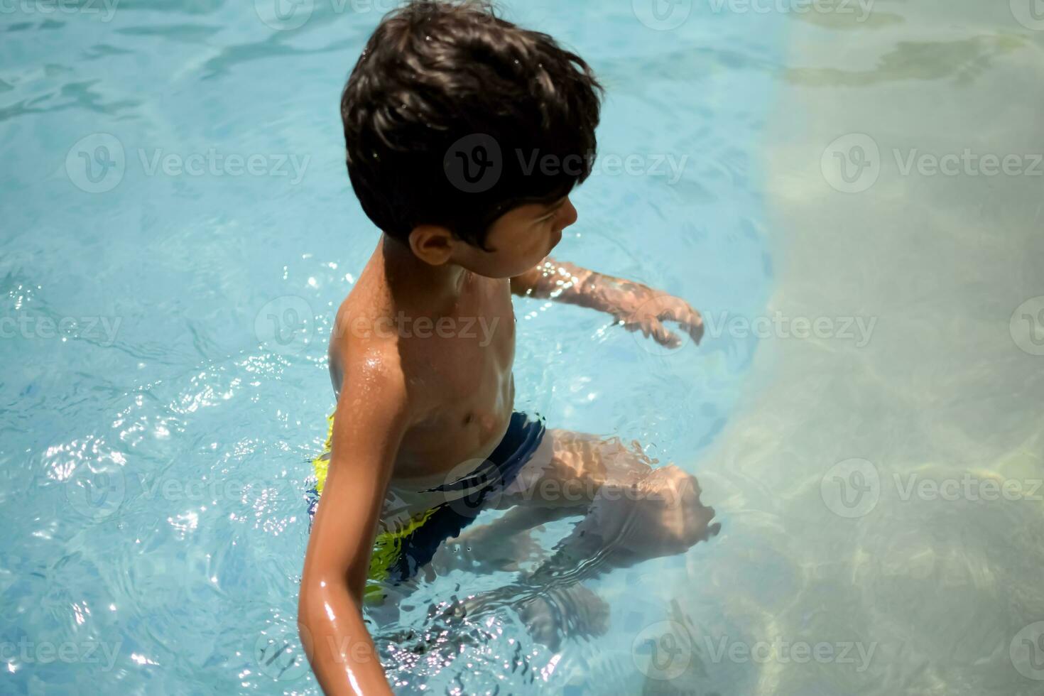 Happy Indian boy swimming in a pool, Kid wearing swimming costume along with air tube during hot summer vacations, Children boy in big swimming pool. photo