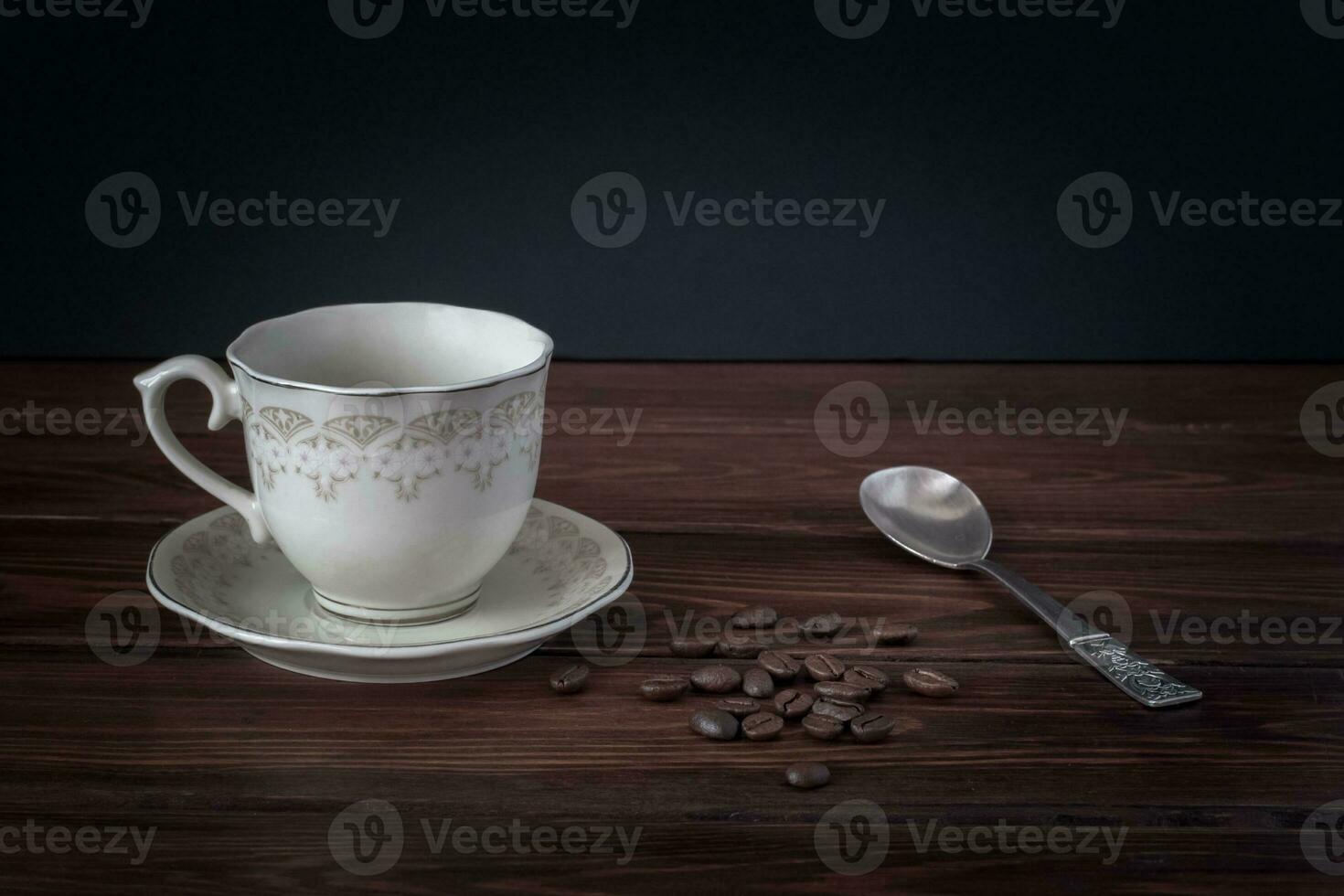 Empty coffee mug on a saucer, coffee beans on dark table boards. Beautiful silver spoon. Place for text. photo