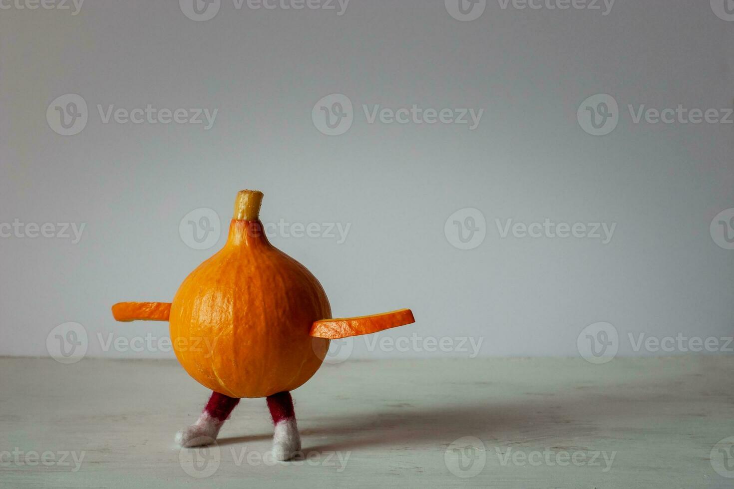 An orange little pumpkin with legs and hands is standing on a white board. Hands are spread out to the sides. The background is light. Copy space. photo