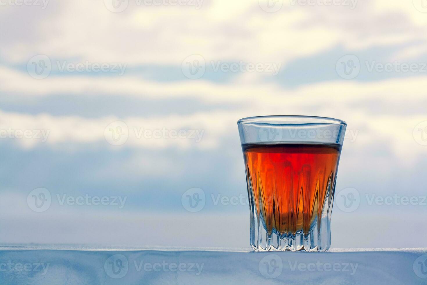 un hermosa vaso de alcohol soportes en un glacial base en contra el cielo. enfriado whisky al aire libre en invierno. naranja bebida en un vaso. Copiar espacio. lado vista. horizontal. foto