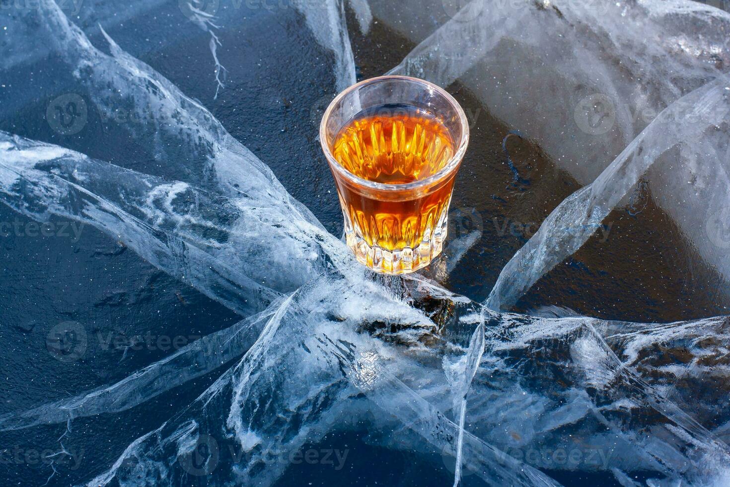 On the cracked ice of the lake is a glass of whiskey. Chilled whiskey. Blue ice with beautiful deep white cracks. Top view from the side. Advertising alcohol products. Horizontal. photo