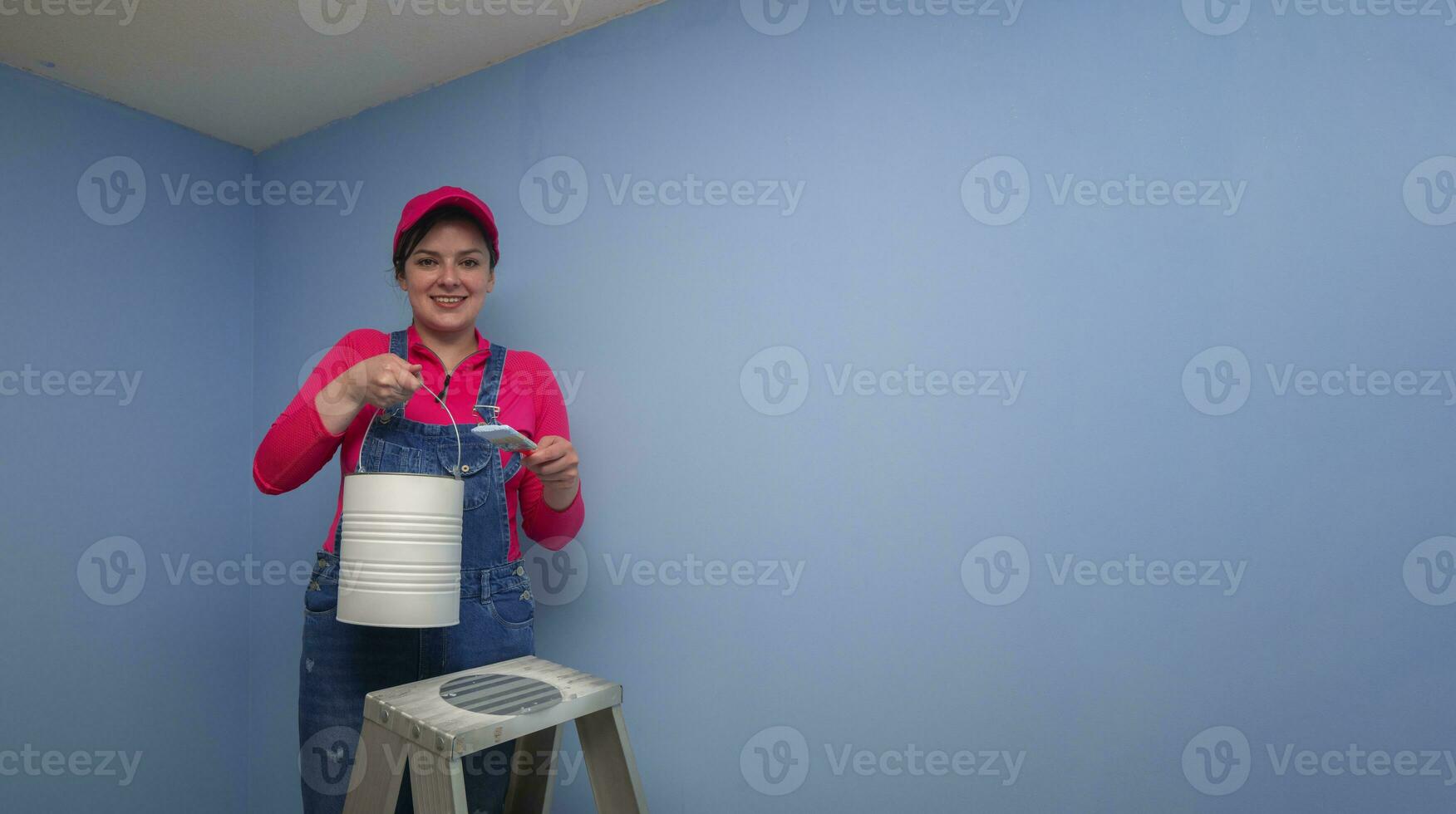 woman dressed in overalls and red t-shirt standing on a metal ladder, seen from the front, holding the brush and the paint jar next to a blue wall photo