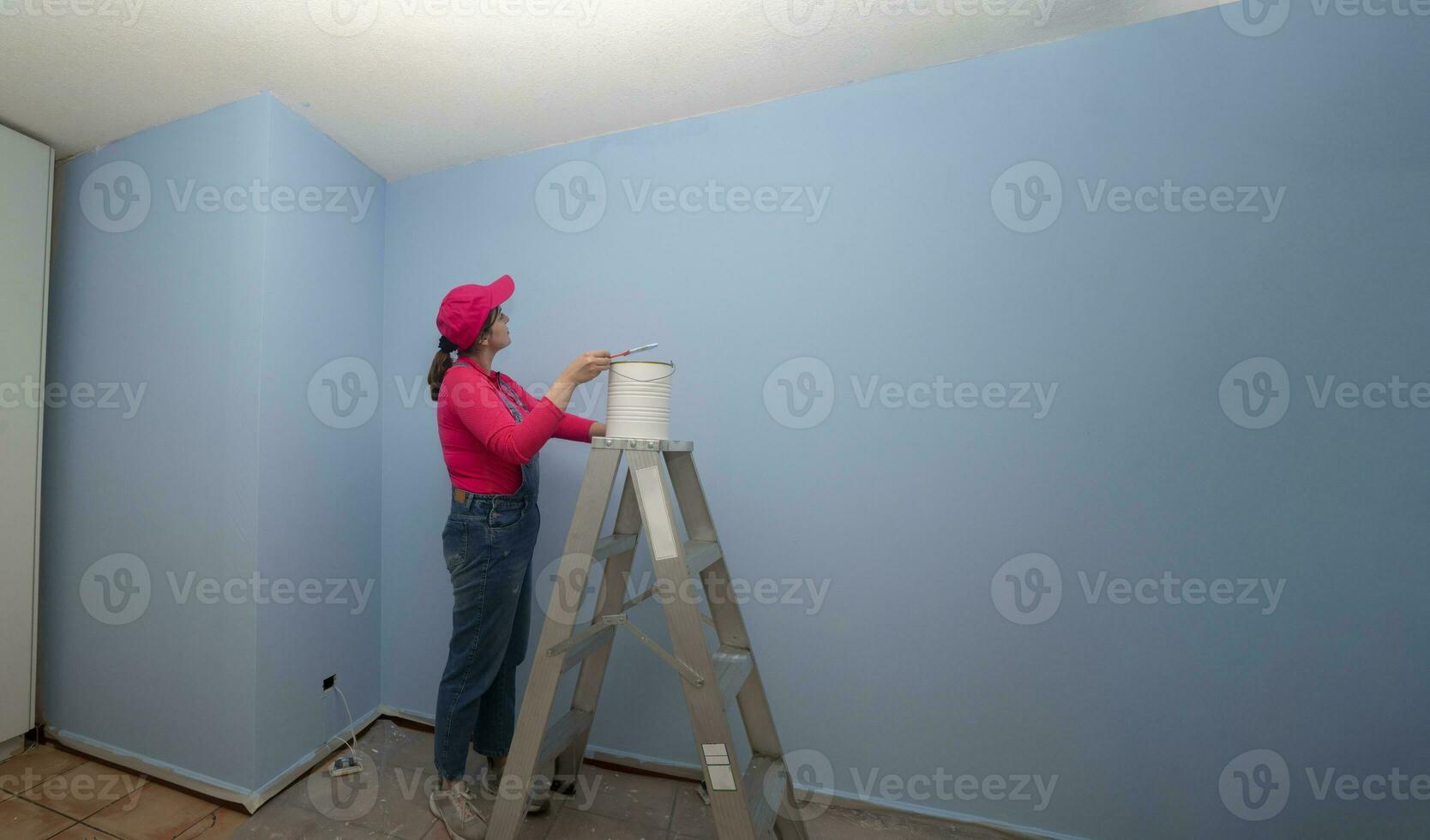 mujer vestido en mono y rojo camiseta yendo arriba un metal escalera con un tarro de pintar siguiente a un azul pared en un vacío habitación foto