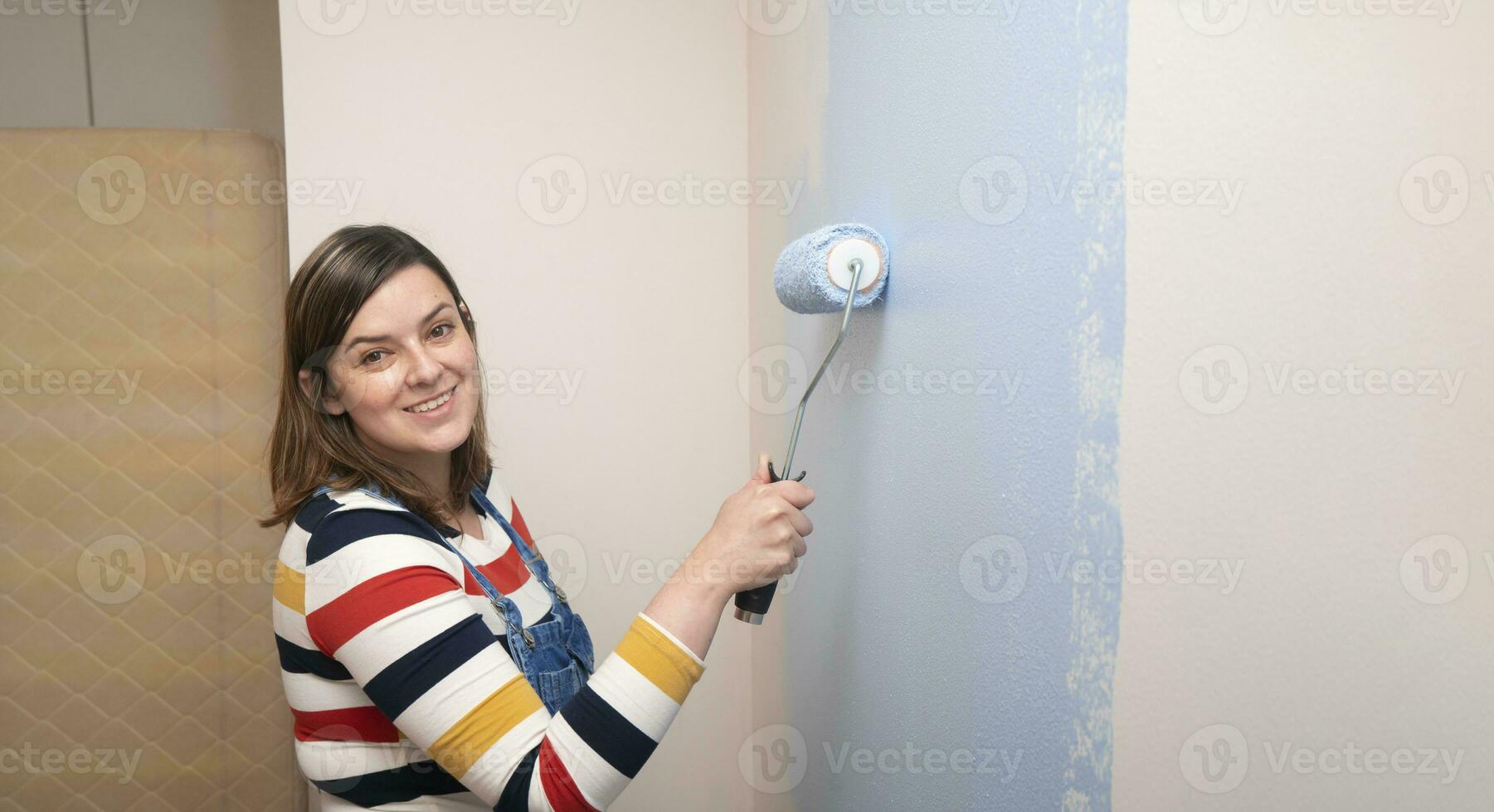 woman standing dressed in overalls and striped blouse, profile view, smiling at camera with a roller in her hand painting a white wall with blue paint photo