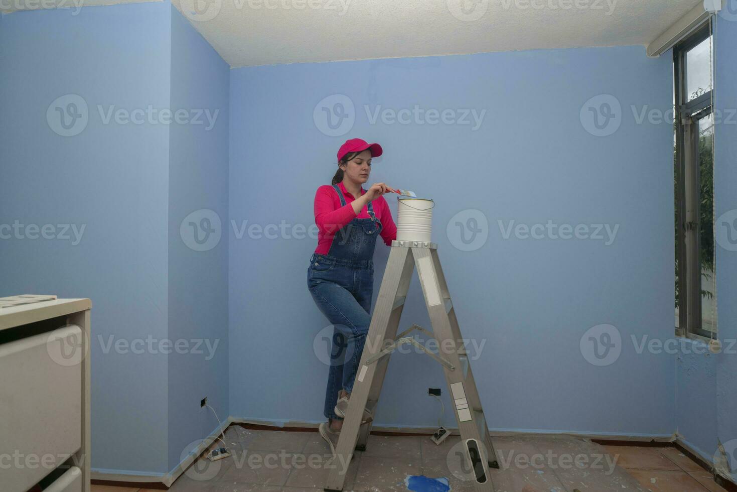 woman dressed in overalls and red t-shirt going up a metal ladder with a jar of paint next to a blue wall in an empty room photo