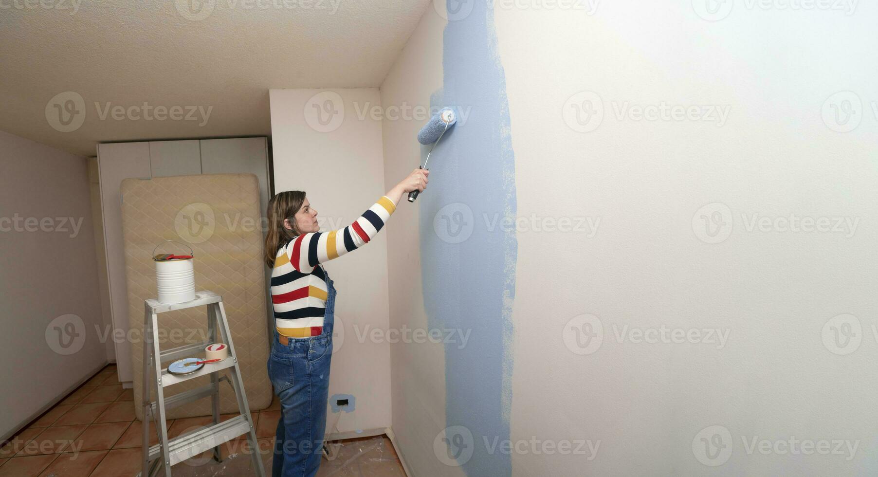 mujer vestido en mono y a rayas blusa, perfil vista, con un rodillo en su mano pintura un blanco pared con azul pintar en un vacío habitación foto