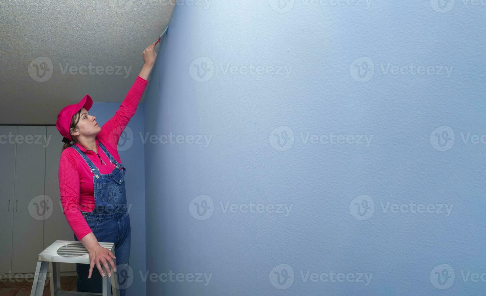 woman dressed in overalls and a red t-shirt on a metal ladder, with a brush in her hand finishing painting a blue wall in an empty room photo
