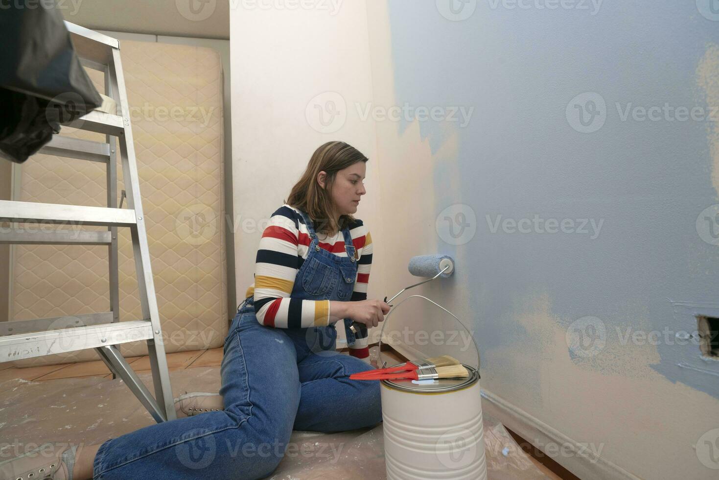 woman dressed in overalls and striped blouse, profile view, painting a white wall with a roller with blue paint, inside an empty room with a metal ladder, paint jar and brushes photo