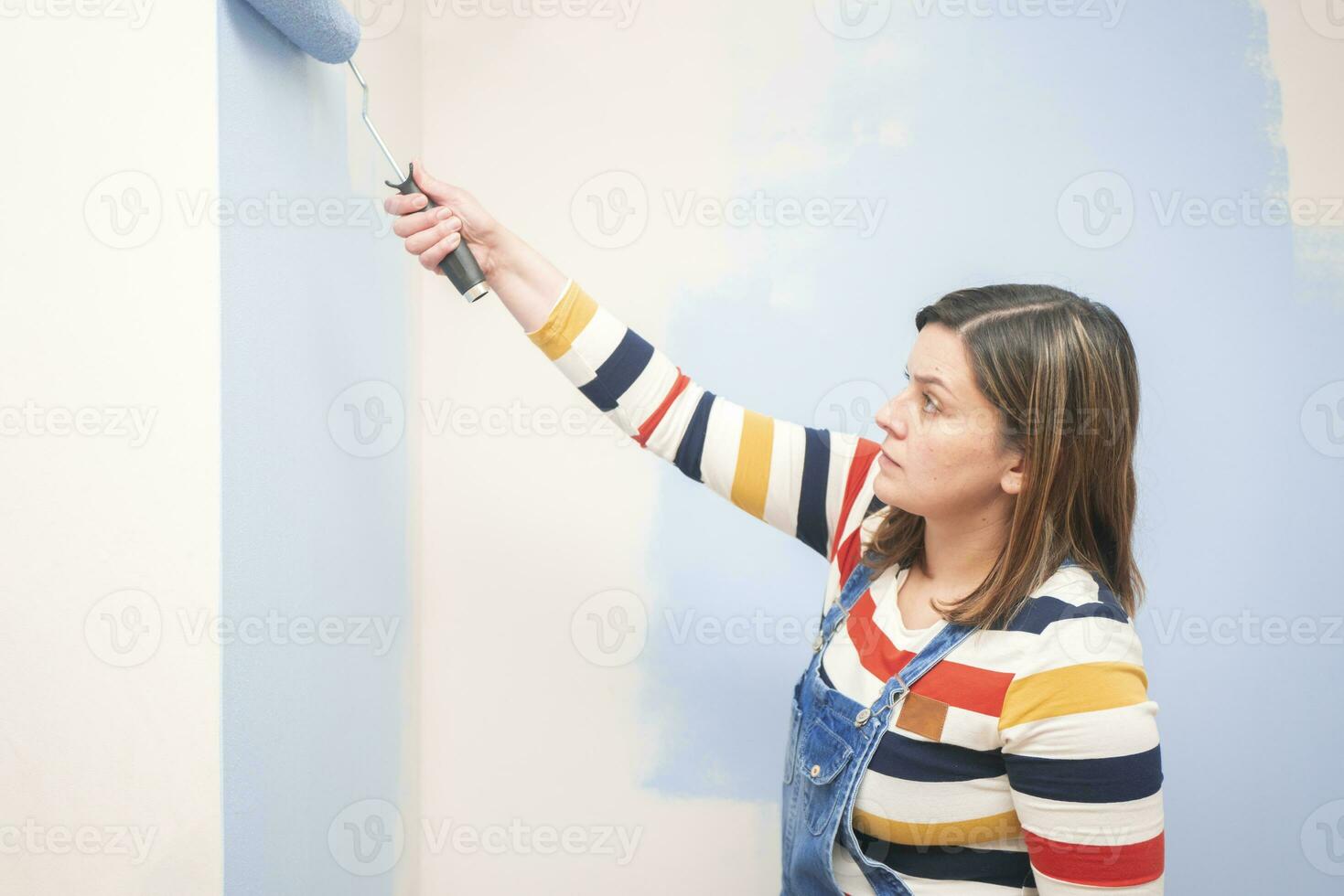 woman standing dressed in overalls and striped blouse, profile view, with a roller in her hand painting a white wall with blue paint photo