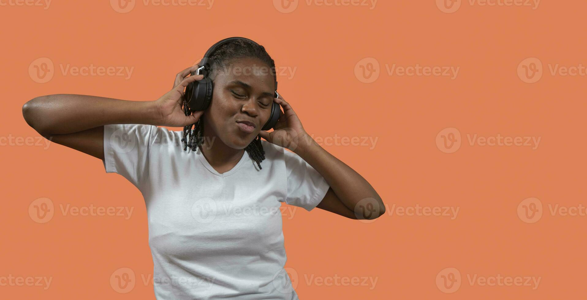 retrato de africano joven mujer escuchando a música en auriculares, ojos cerrado y participación auriculares con manos en contra naranja de colores antecedentes foto