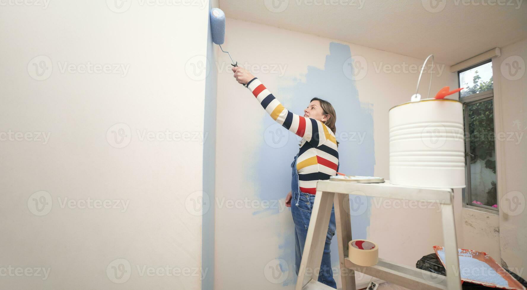 jar of paint on a ladder with a background of a standing woman dressed in overalls and striped blouse, seen from behind, painting a white wall with a roller with blue paint photo