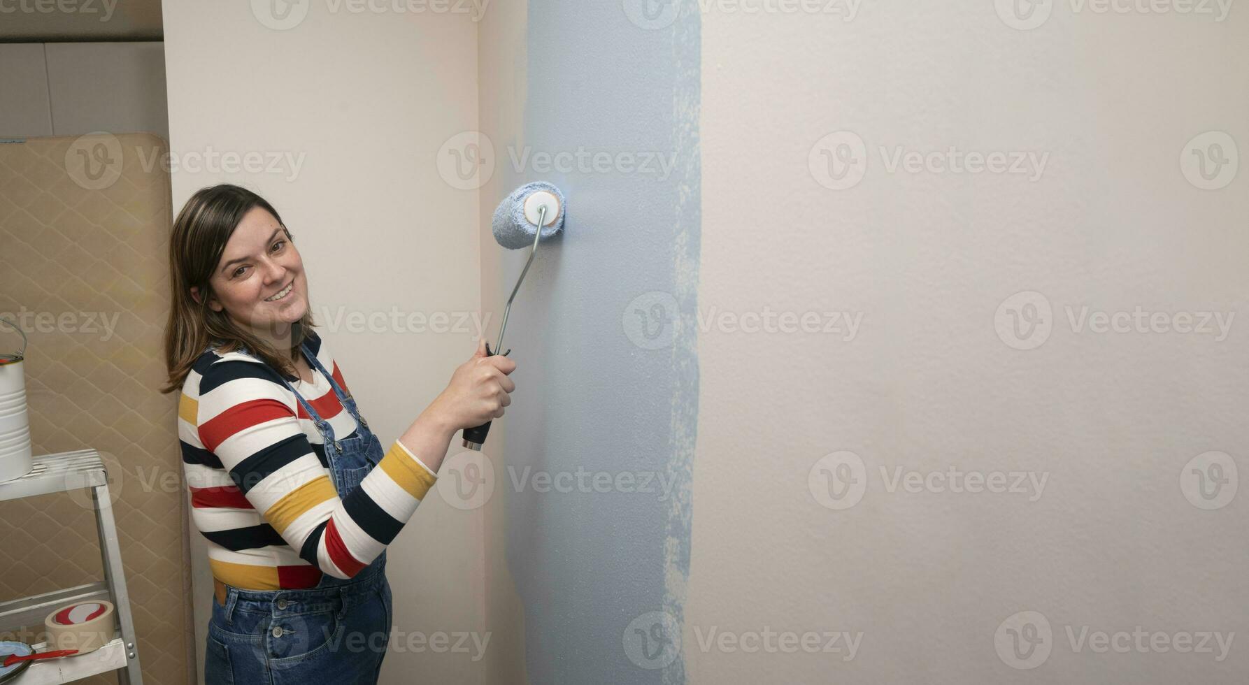 woman standing dressed in overalls and striped blouse, profile view, smiling at camera with a roller in her hand painting a white wall with blue paint photo