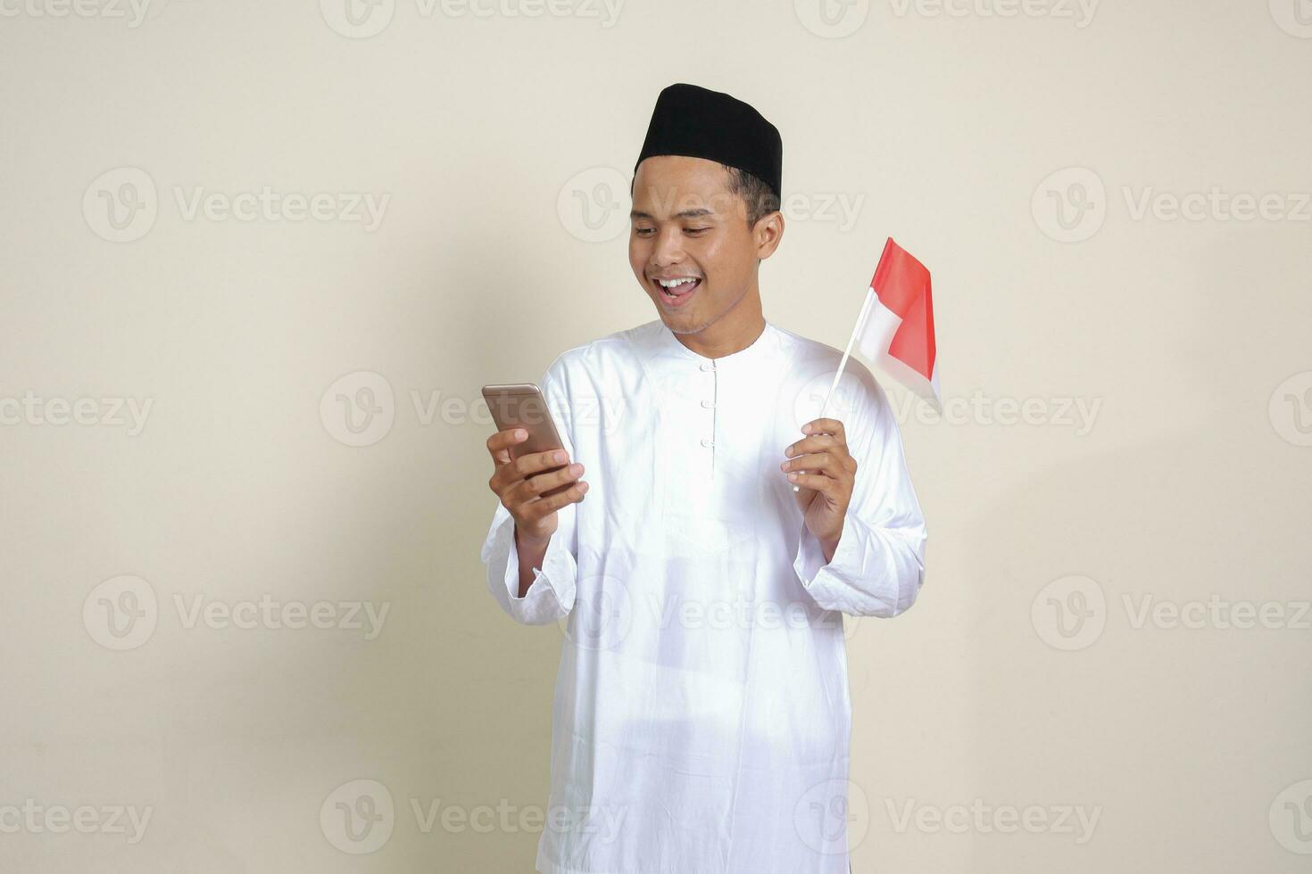 Portrait of attractive Asian muslim man in white shirt with skullcap holding indonesia flag while showing mobile phone. Isolated image on gray background photo