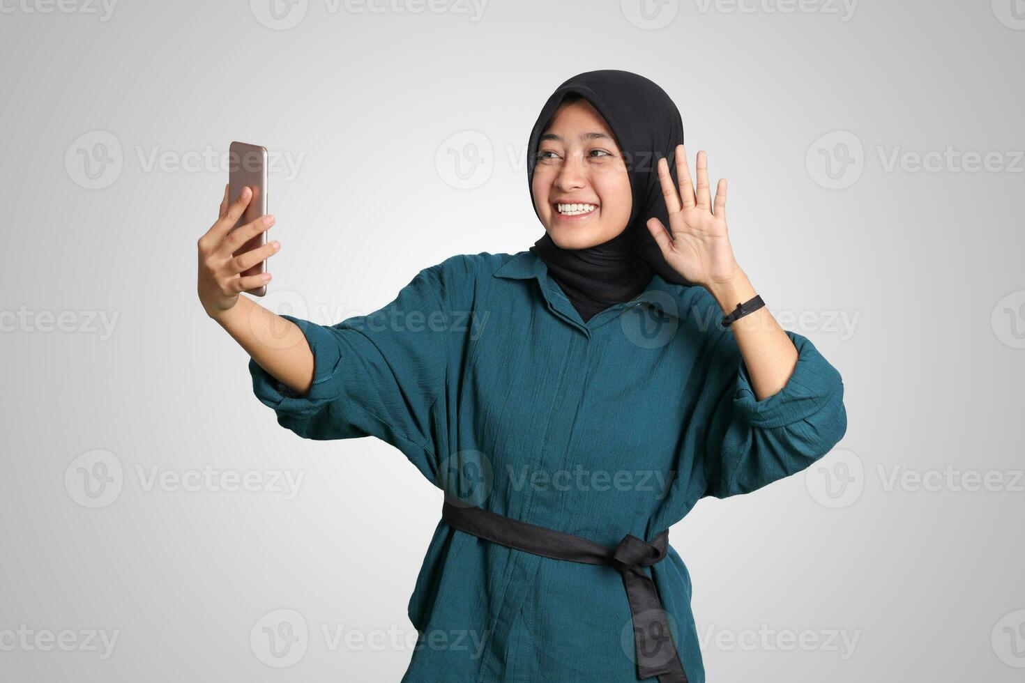 Portrait of excited Asian muslim woman with hijab holding mobile phone while in video calling with her hand gesture saying hi. Advertising concept. Isolated image on white background photo