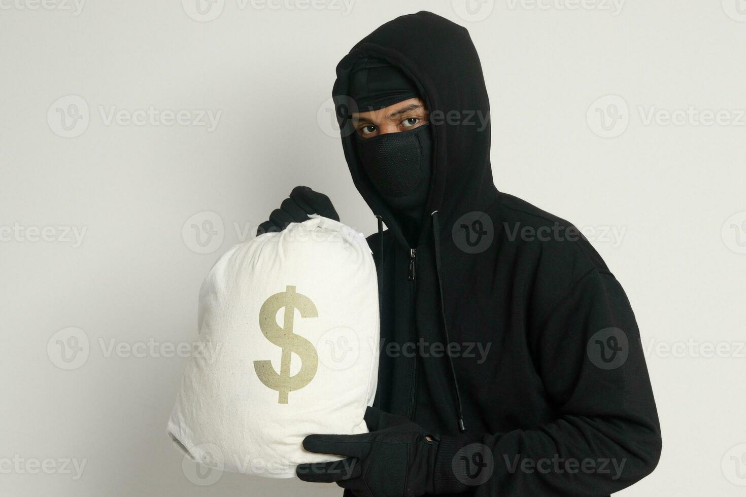 misterioso ladrón ladrón hombre vistiendo negro capucha y máscara que lleva un bolso lleno de dinero. aislado imagen en gris antecedentes foto