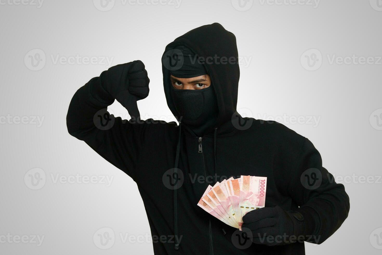 Portrait of mysterious man wearing black hoodie and mask stealing one hundred thousand rupiah successfully from victim. Isolated image on gray background photo