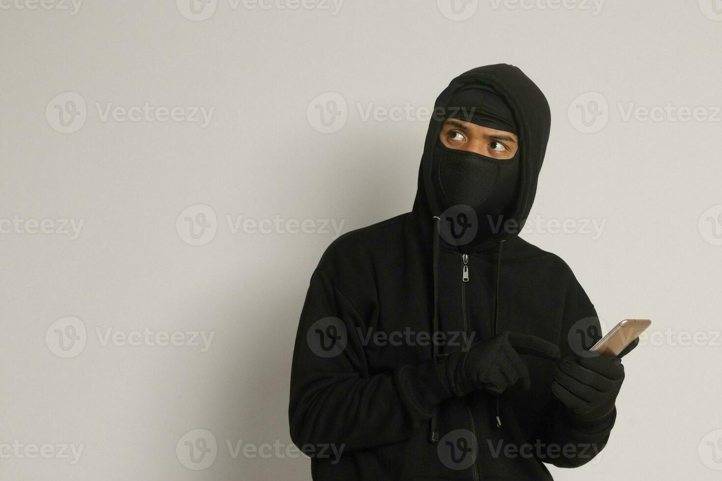 Portrait of mysterious man wearing black hoodie and mask doing hacking activity on mobile phone, hacker holding a smartphone. Isolated image on gray background photo