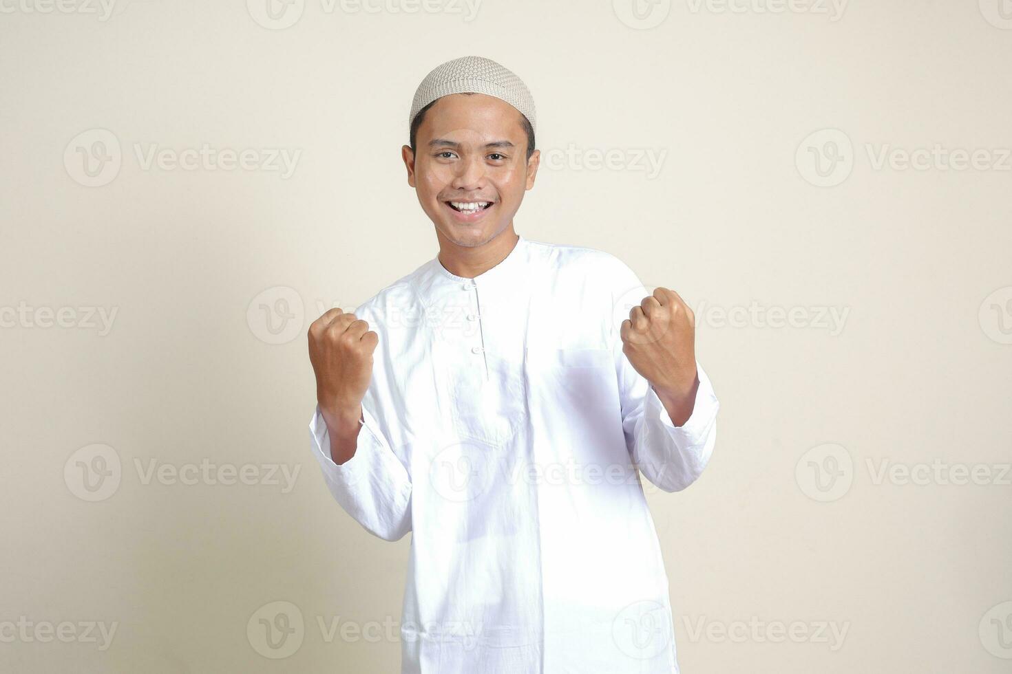 Portrait of attractive Asian muslim man in white shirt raising his fist, celebrating success. Isolated image on gray background photo