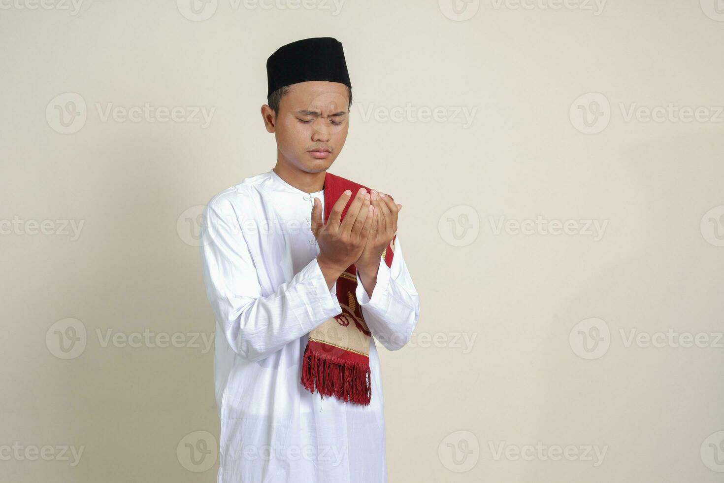 Portrait of attractive Asian muslim man in white shirt with skullcap praying earnestly with his hands raised. Isolated image on gray background photo