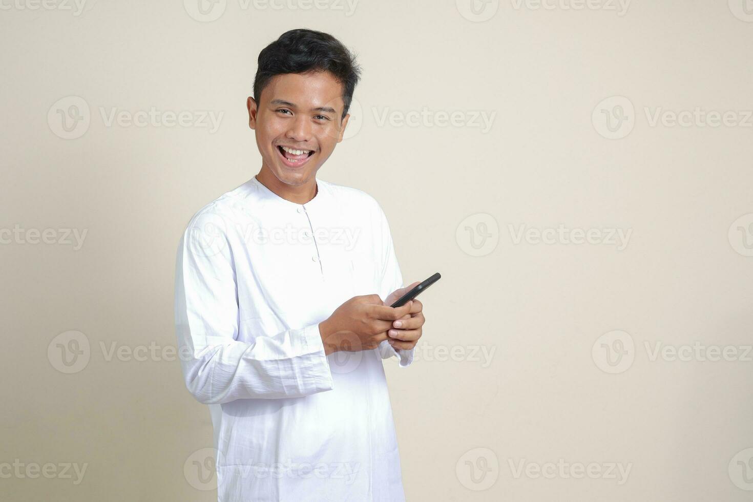 Portrait of attractive Asian muslim man in white shirt holding mobile phone with smiling expression on face. Advertising concept. Isolated image on gray photo
