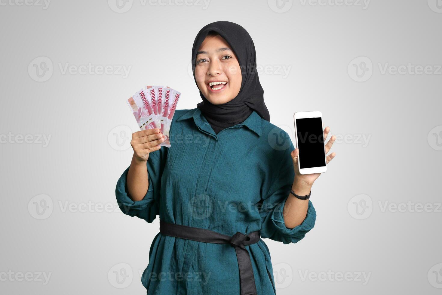 Portrait of excited Asian muslim woman with hijab, showing one hundred thousand rupiah while showing blank screen mockup mobile phone. Financial and savings concept. Isolated image on white background photo