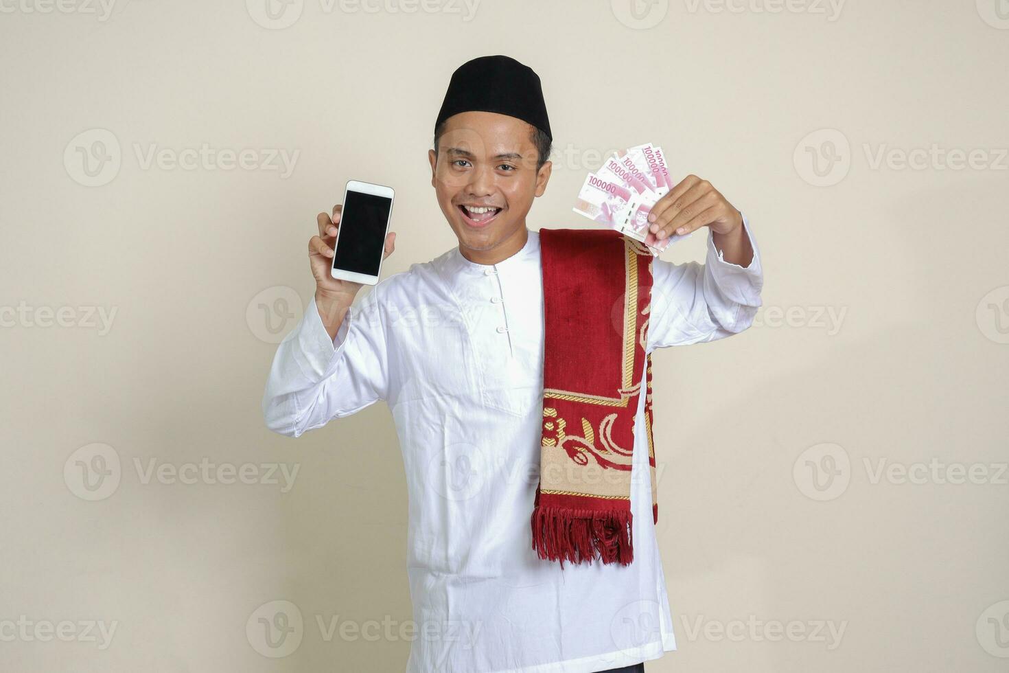 Portrait of attractive Asian muslim man in white shirt with skullcap showing one hundred thousand rupiah while showing blank screen mobile phone. Financial and shopping concept. Isolated image on gray photo