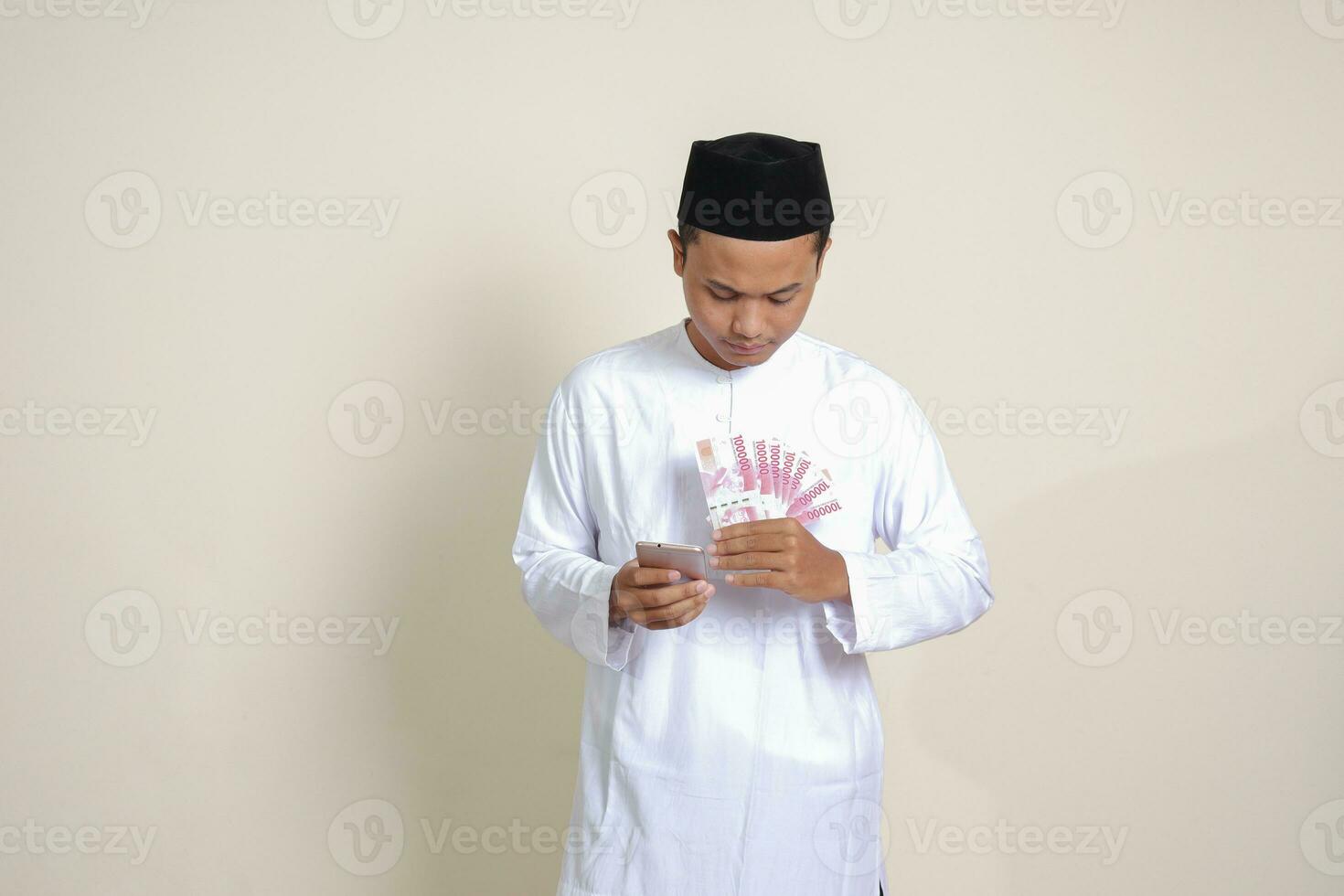 Portrait of attractive Asian muslim man in white shirt showing one hundred thousand rupiah while using mobile phone. Financial and savings concept. Isolated image on gray background photo