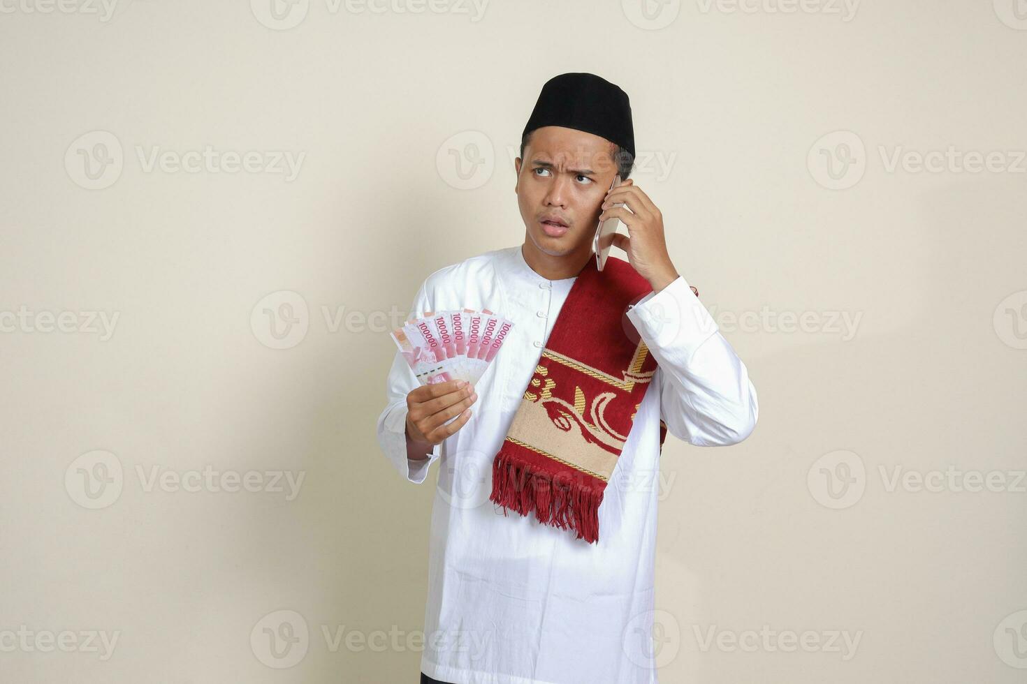Portrait of attractive Asian muslim man in white shirt showing one hundred thousand rupiah while using mobile phone. Financial and savings concept. Isolated image on gray background photo