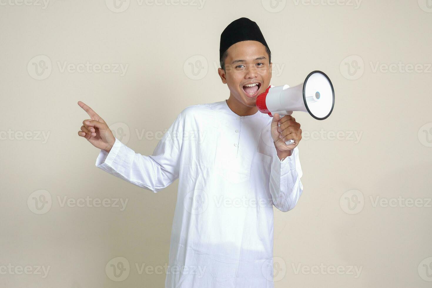 Portrait of attractive Asian muslim man in white shirt with skullcap speaking louder using megaphone, promoting product. Advertising concept. Isolated image on grey background photo