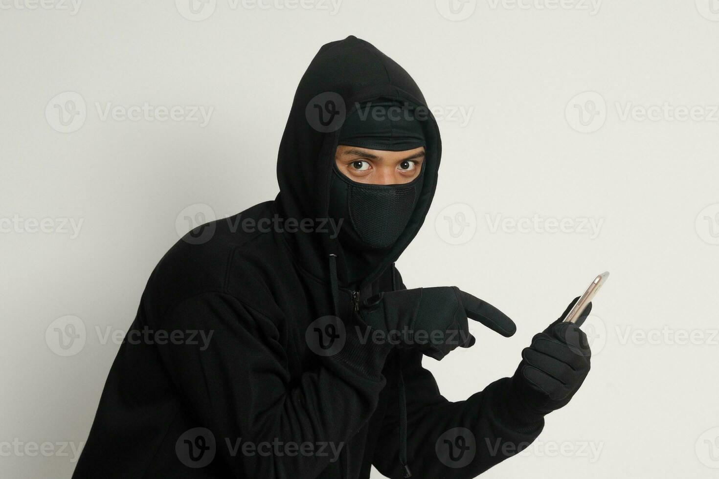 Portrait of mysterious man wearing black hoodie and mask doing hacking activity on mobile phone, hacker holding a smartphone. Isolated image on gray background photo