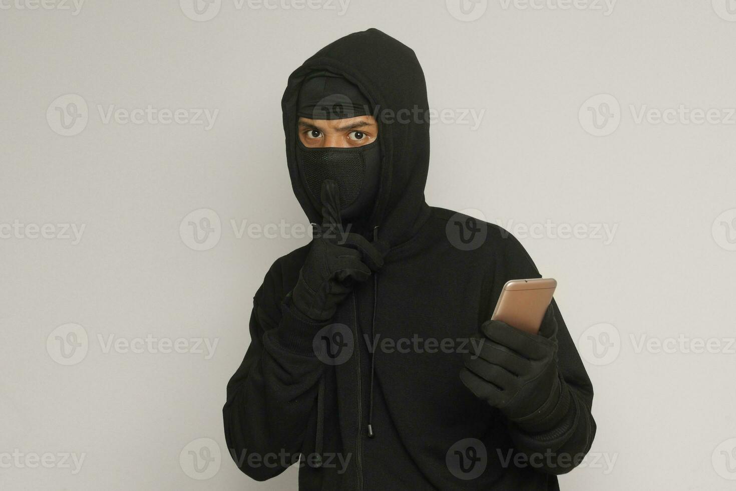 Portrait of mysterious man wearing black hoodie and mask doing hacking activity on mobile phone, hacker holding a smartphone. Isolated image on gray background photo
