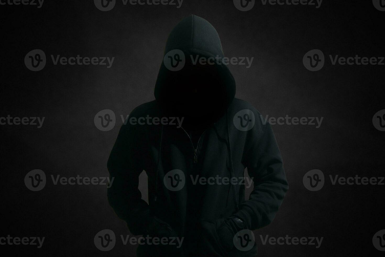 Mysterious man wearing black hoodie standing against dark background. Hacker, crime, and cyber security concept. photo