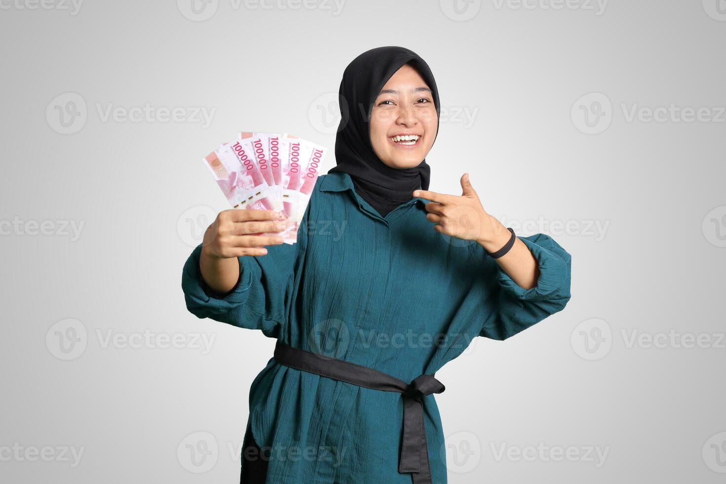 Portrait of excited Asian hijab woman in casual outfit showing and pointing one hundred thousand rupiah. Financial and savings concept. Isolated image on white background photo