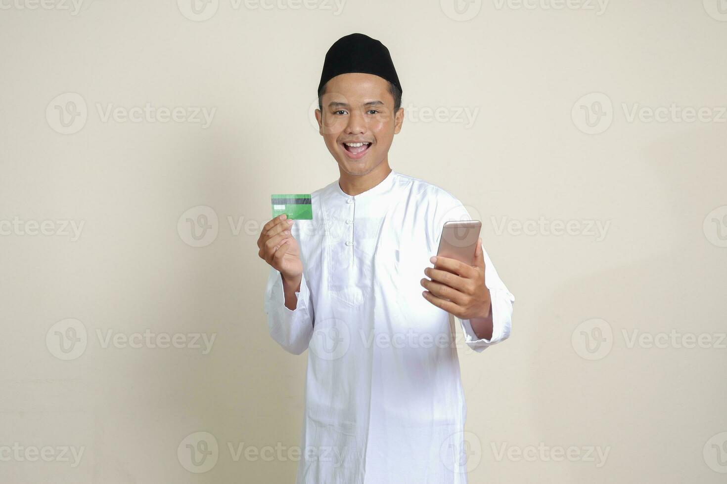 Portrait of attractive Asian muslim man in white shirt with skullcap holding a mobile phone and presenting credit card. Isolated image on gray background photo