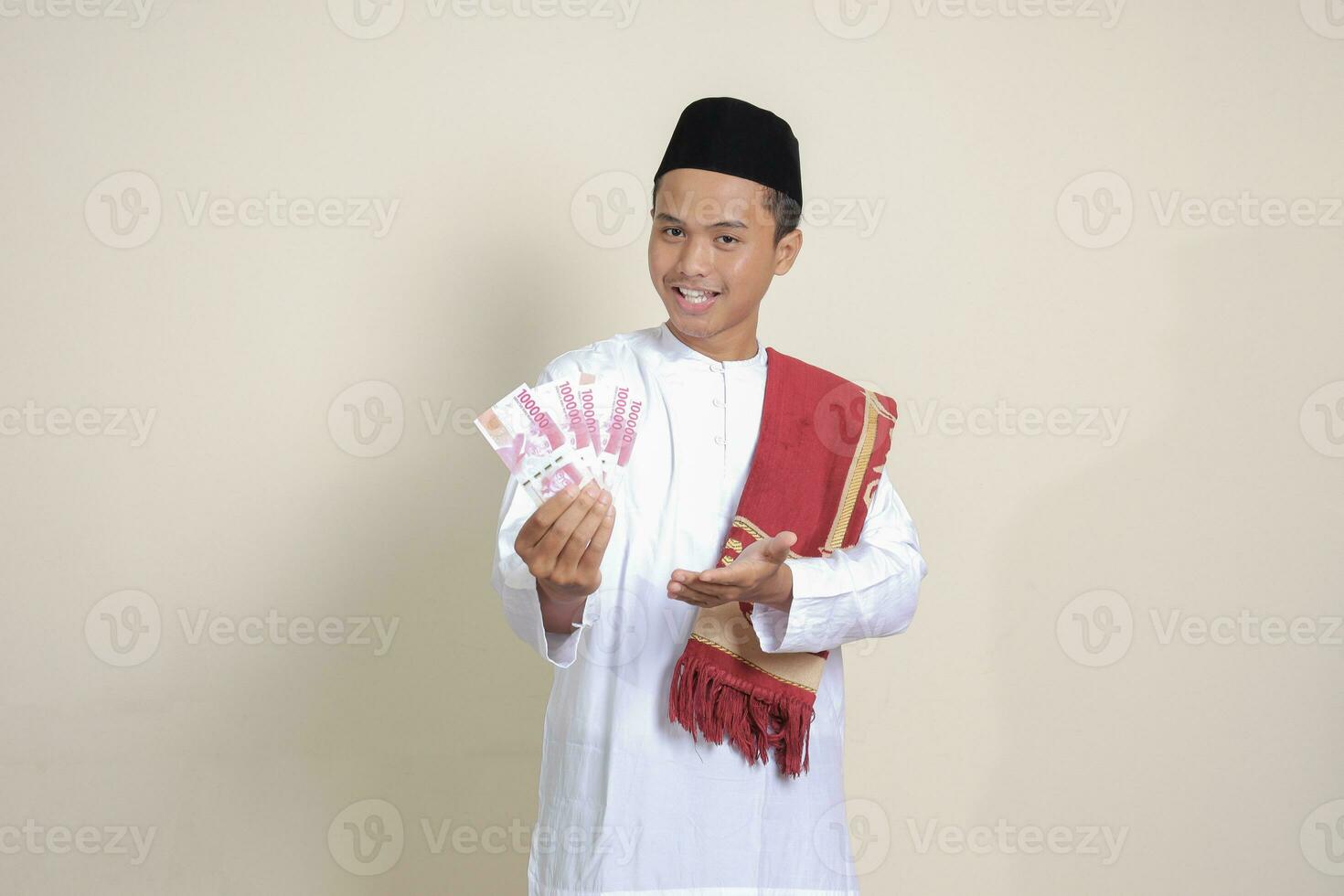 Portrait of attractive Asian muslim man in white shirt showing one hundred thousand rupiah while pointing to the side. Financial and savings concept. Isolated image on gray background photo