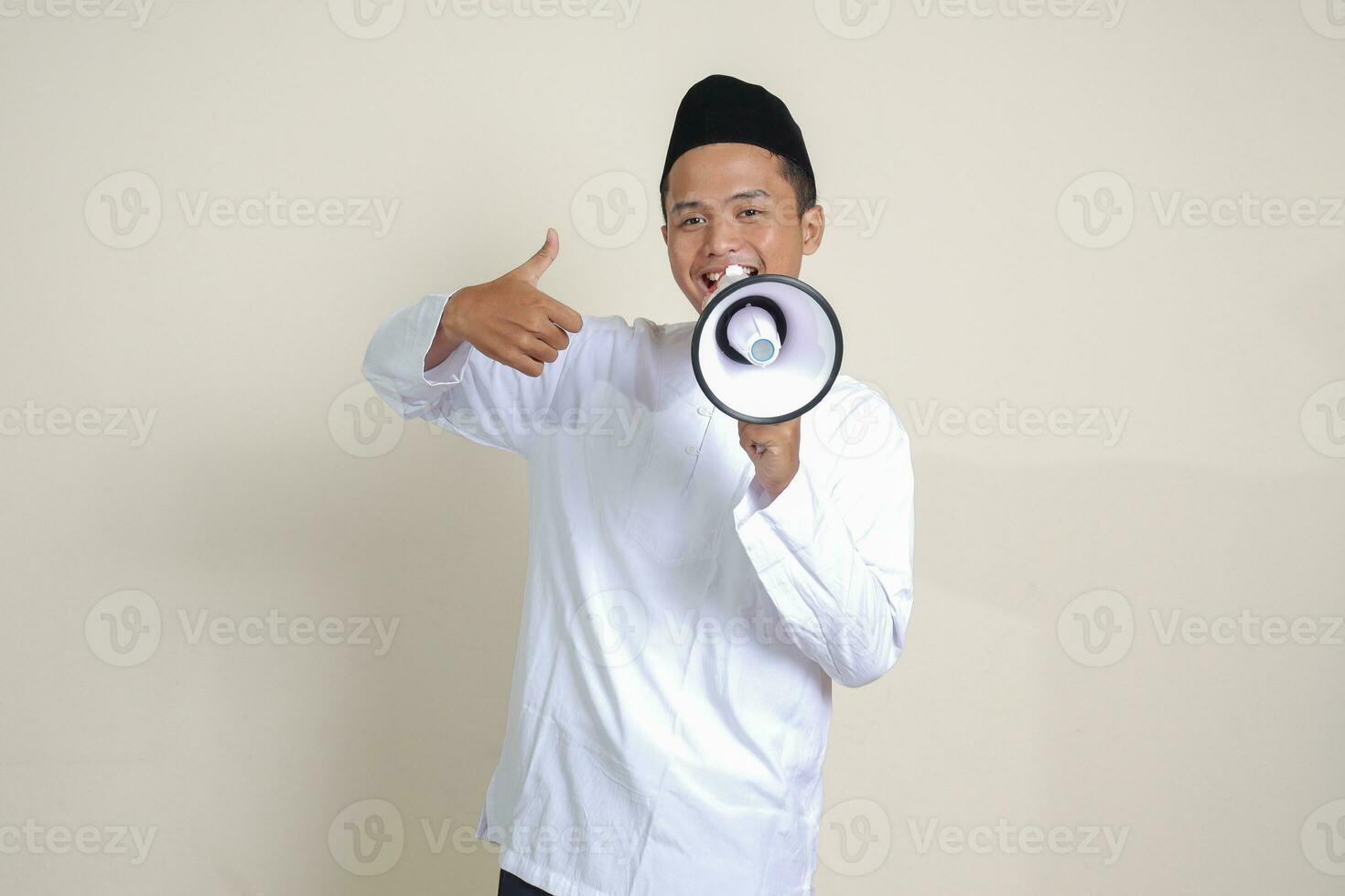 Portrait of attractive Asian muslim man in white shirt with skullcap speaking louder using megaphone, promoting product. Advertising concept. Isolated image on grey background photo