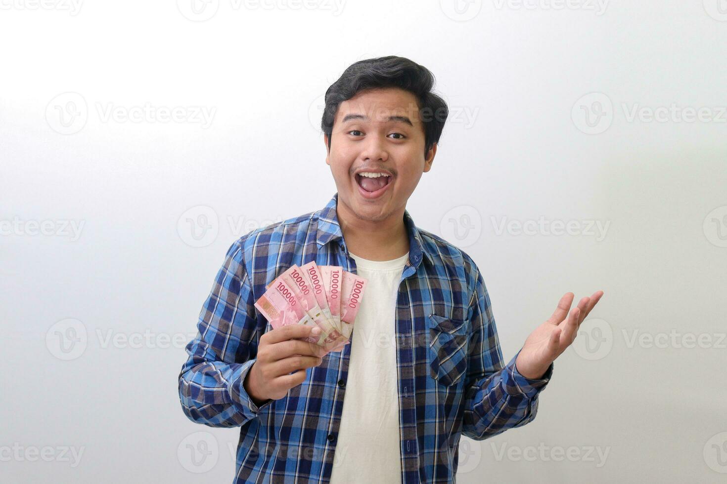 retrato de emocionado asiático hombre en azul tartán camisa en pie en contra blanco fondo, demostración uno cien mil rupia mientras señalando a el lado. financiero y ahorros concepto. foto