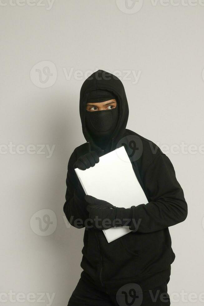 Mysterious robber thief man wearing black hoodie and mask stealing laptop and sneak out. Isolated image on gray background photo