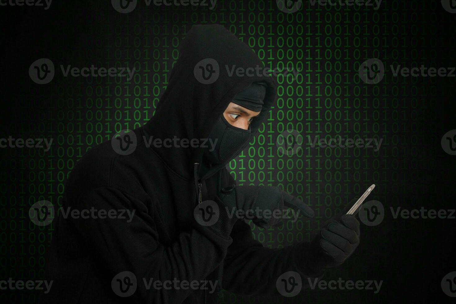 Portrait of mysterious man wearing black hoodie and mask doing hacking activity on mobile phone, hacker holding a smartphone. Isolated image on gray background photo